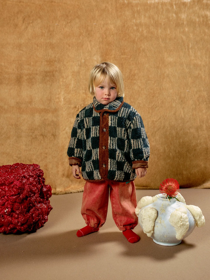 A young child stands against a textured tan backdrop wearing a dark green and cream checkered jacket with brown trim, loose rust-red pants, and red socks. Decorative red and white sculptural objects sit on the floor beside the child.