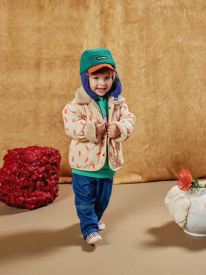A toddler wearing a cream puffer jacket with orange leaf prints, blue jeans, a green and orange cap, and a blue hooded neck warmer, standing against a textured gold backdrop with decorative props.
