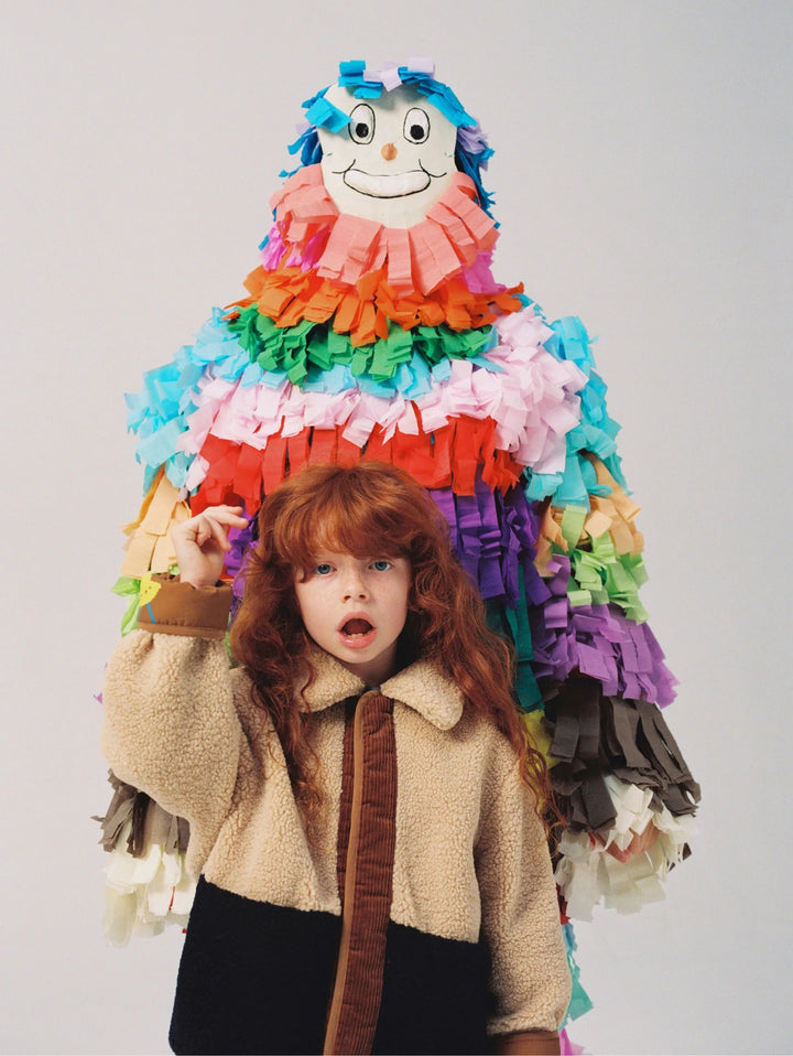 A young child with long red hair stands in front of a large, colorful paper piñata shaped like a smiling character, wearing a beige sherpa jacket with a brown collar and dark skirt, against a plain light gray background.