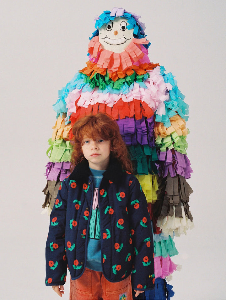 A child with curly red hair stands in front of a large, colorful paper-tear piñata figure with a smiling face. The child is wearing a navy blue quilted jacket with a red and green cherry print, a teal shirt, and orange pants, against a plain light gray studio background.