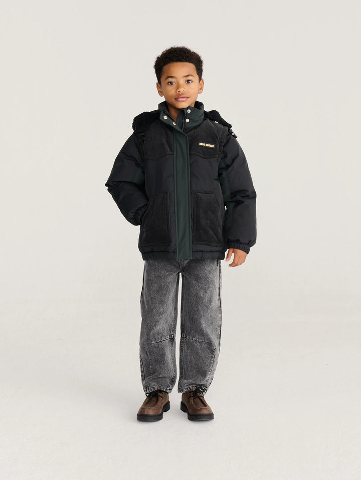 A young boy modeling a black puffer jacket with a dark green inner collar, paired with gray washed jeans and brown shoes, standing against a light gray studio background.