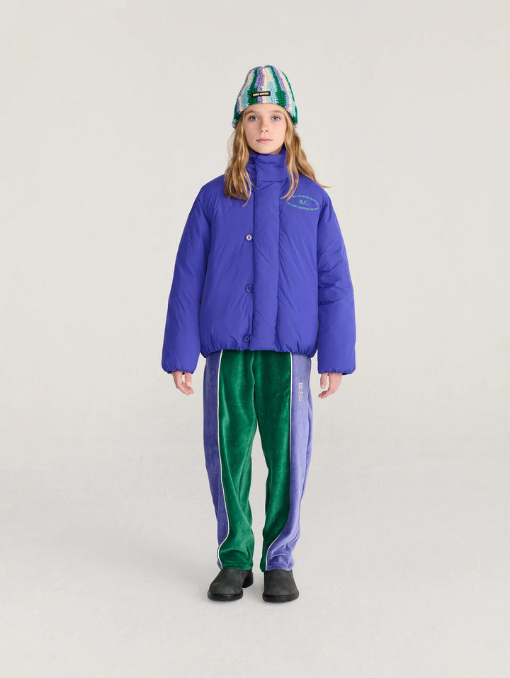 A child model wearing a bright blue quilted puffer jacket, green velvet track pants with purple side panels, and a green and blue patterned beanie, standing against a light gray studio background.