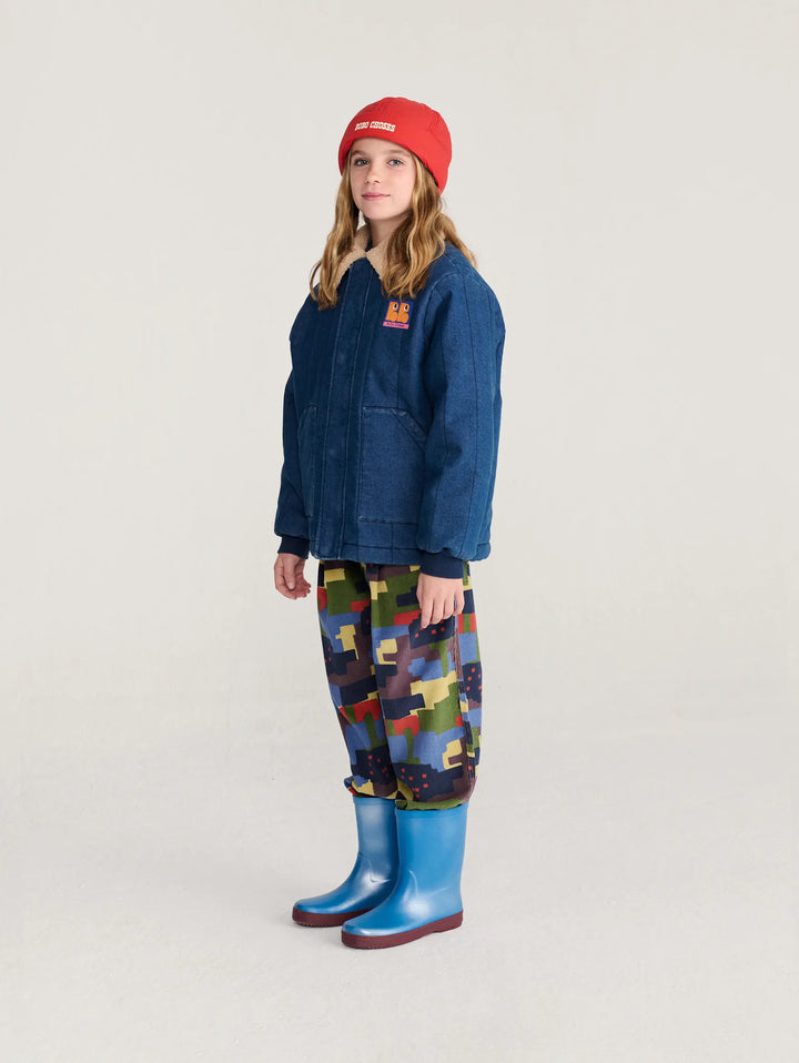 A child model wearing a dark blue denim jacket, a red knit beanie, a colorful geometric print pants, and bright blue rain boots, standing against a plain light gray studio background.