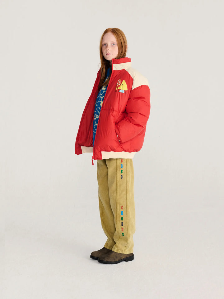 A model stands in profile wearing a bright red puffer jacket with cream panels, tan pants with colorful button details, and dark brown boots, against a light gray studio background.