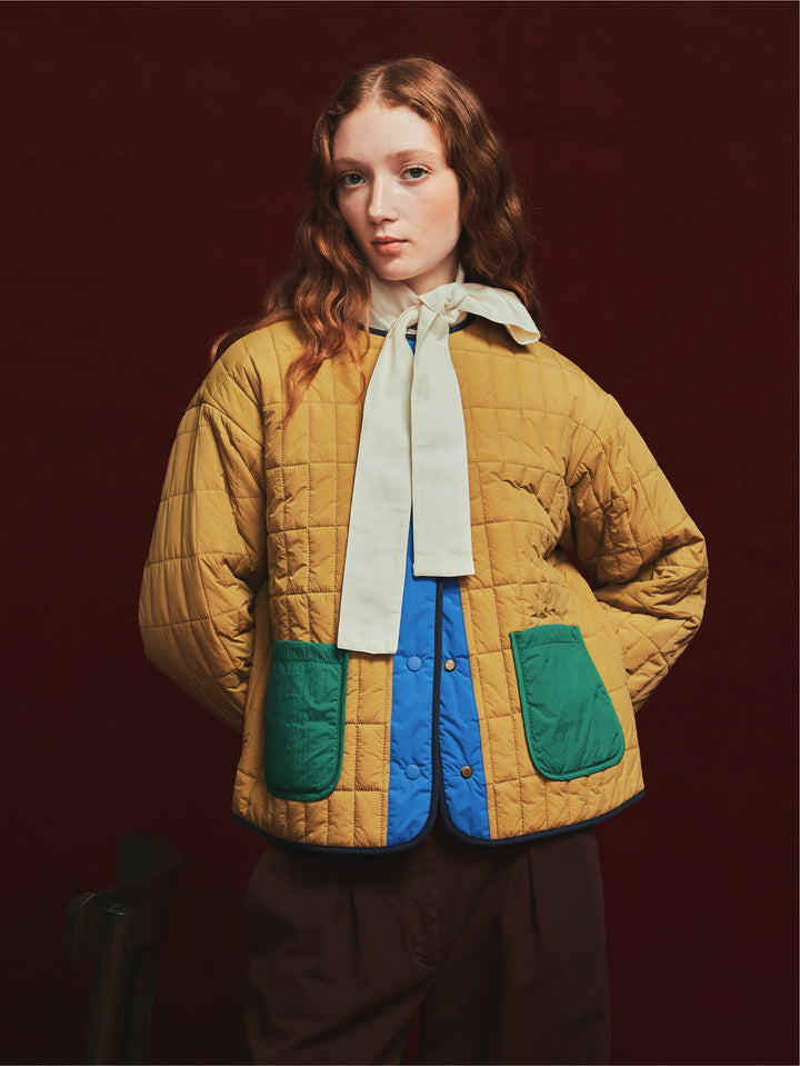 A model wearing a mustard yellow quilted jacket with green pockets, a cream bow tie, and a blue shirt, set against a dark reddish-brown studio background.