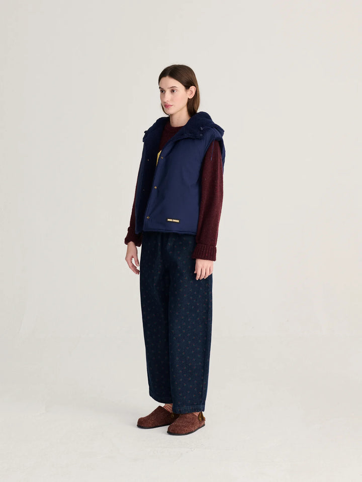 A model stands in profile wearing a navy blue sleeveless puffer vest over a burgundy sweater, paired with dark patterned wide-leg trousers and brown suede shoes, against a plain light gray studio background.