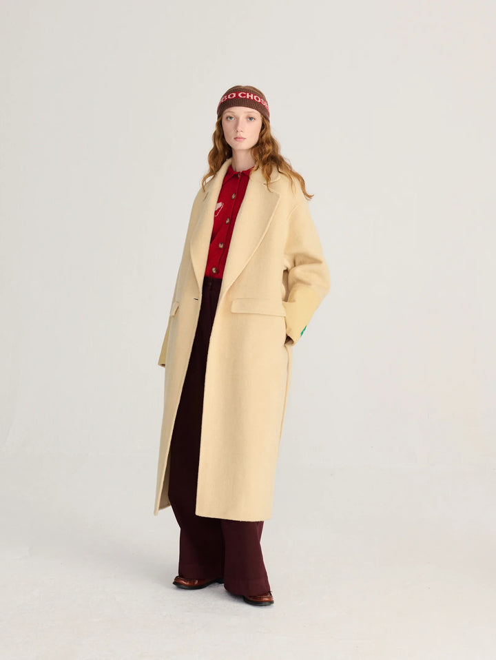 A model stands against a light gray studio background wearing a long cream-colored overcoat over a red patterned top and dark burgundy pants, with a burgundy beanie featuring white text.