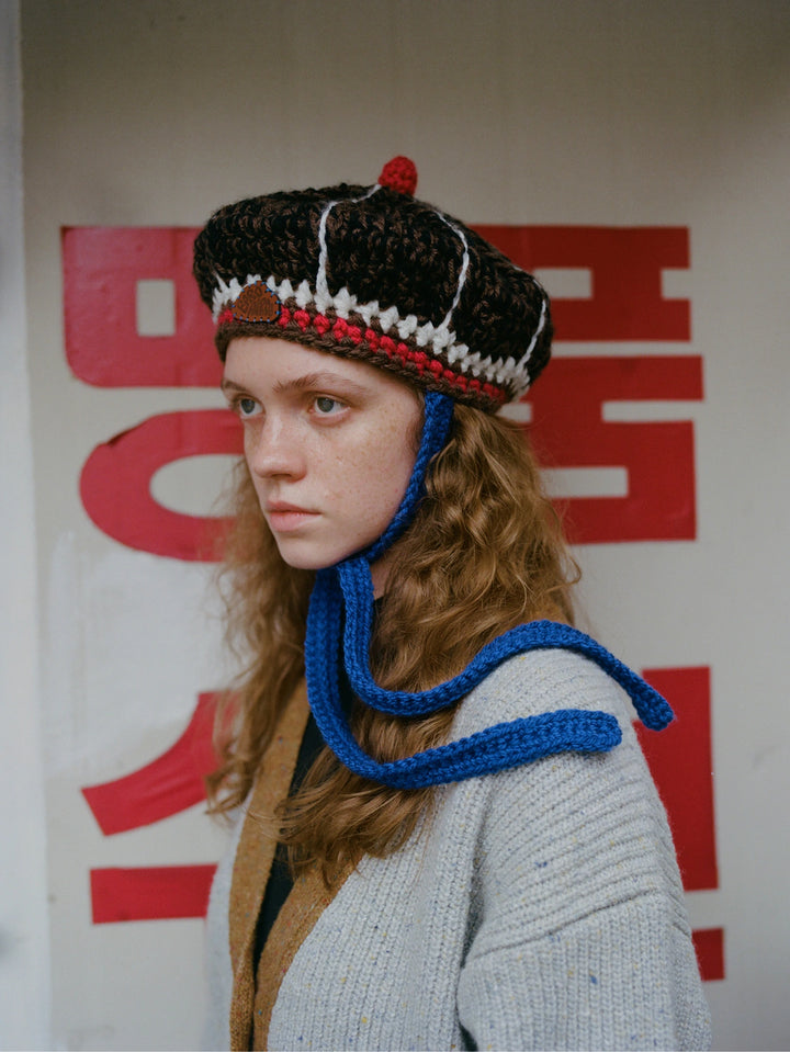 A person wearing a crocheted black, white, and red beret-style hat with a long blue chin strap, paired with a light gray knit cardigan, standing in front of a white wall with large red Korean characters.