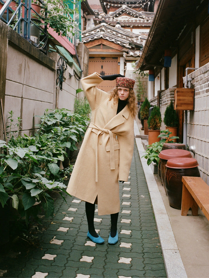 A woman stands in a narrow outdoor alley wearing a long beige trench coat with a waist tie, a patterned brown hat, black tights, and bright blue shoes. The setting includes potted plants, brick paving, and traditional-style buildings.