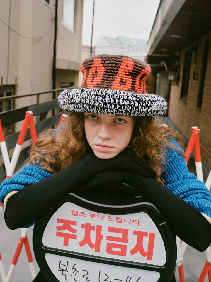 Model wearing a brown crochet bucket hat with orange logo lettering and houndstooth brim, black scarf and blue knit sweater, holding a Korean no-parking sign