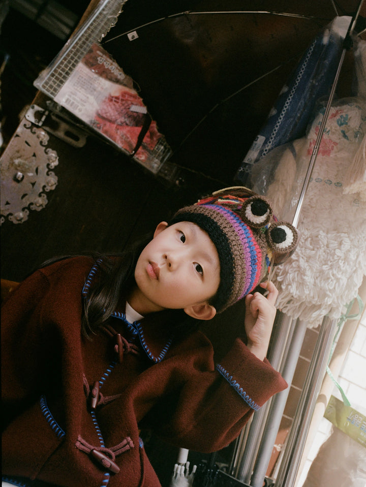 A young girl wearing a brown coat with blue trim and a colorful knit hat with owl ears, looking up in a shop setting.