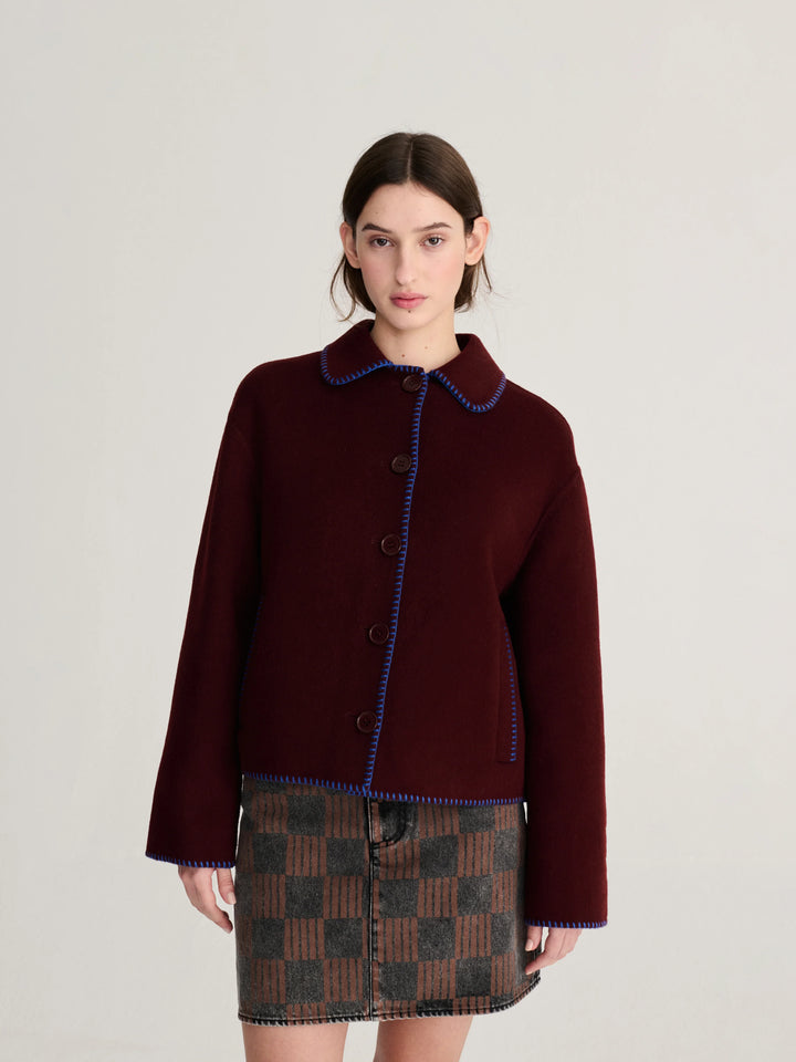 A model wearing a dark burgundy cropped jacket with blue trim and a brown and black checkered mini skirt, photographed against a neutral light gray background.