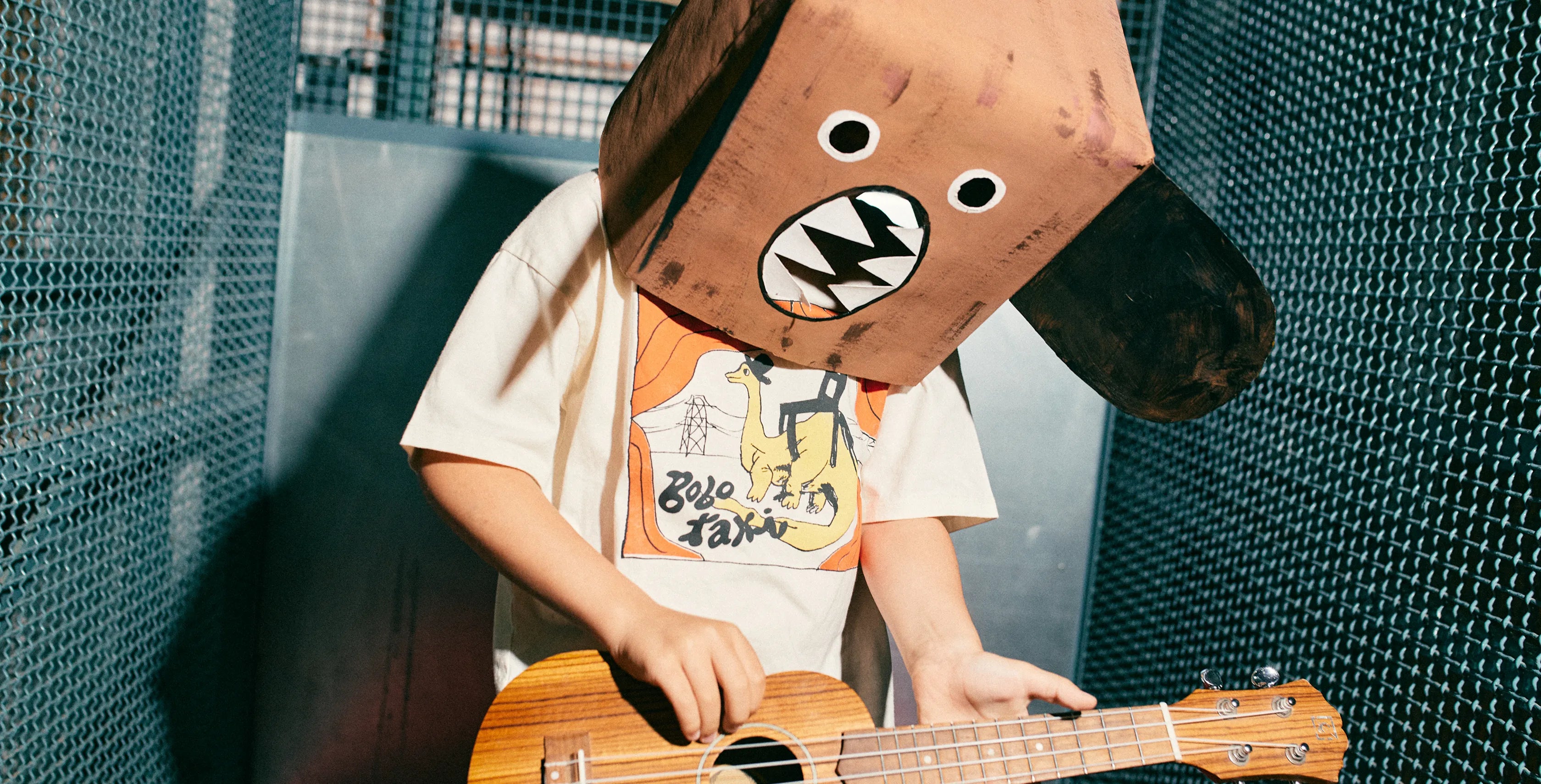 A kid wearing a large brown paper box head with cartoon eyes and a black mouth, playing a wooden ukulele in front of a dark mesh screen. The person is wearing a white graphic t-shirt.