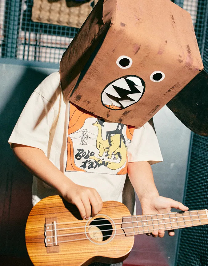 A kid wearing a large brown cardboard box head with cartoon eyes and a mouth, playing a wooden ukulele. The person is wearing a white graphic t-shirt with a yellow dinosaur print.