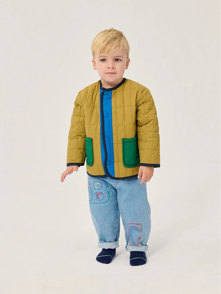 A young blonde boy stands against a light gray studio background wearing a quilted mustard yellow jacket with green pockets over a blue zip-up shirt, light blue jeans with colorful embroidered letters, and navy socks.