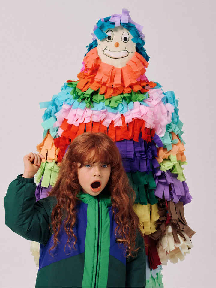A child with long curly red hair stands in front of a large, colorful paper piñata shaped like a clown, wearing a green and blue puffer jacket against a plain light gray background.