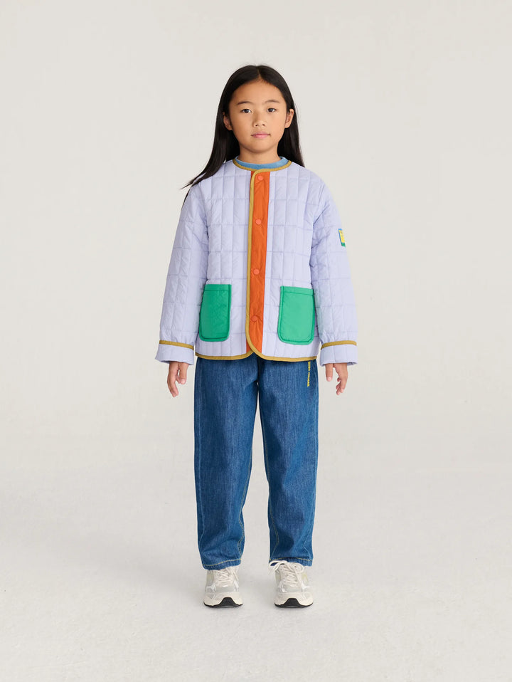 A young girl stands against a light gray studio background wearing a light lavender quilted jacket with bright green pockets, a bright orange front placket, and gold trim, paired with blue jeans and white sneakers.