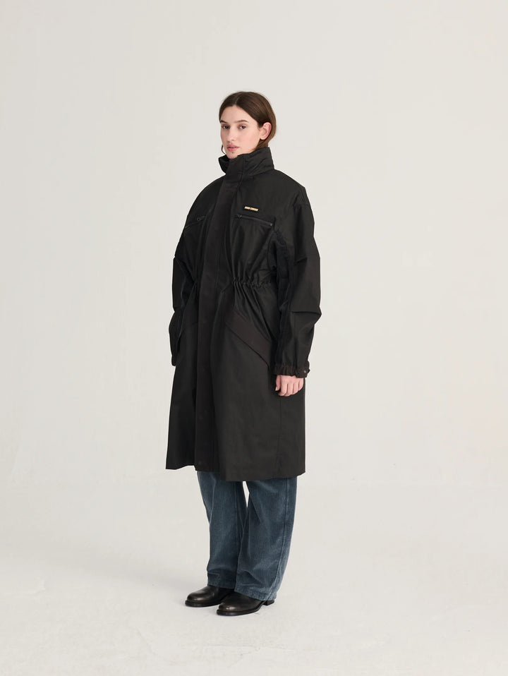 A model wearing a long black coat with a high collar and drawstring waist, paired with blue jeans and black shoes, standing against a light gray studio background.