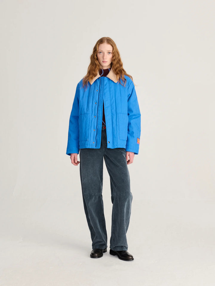 A model stands against a light gray studio background wearing a bright blue quilted jacket with a cream collar, dark gray corduroy pants, and black shoes.