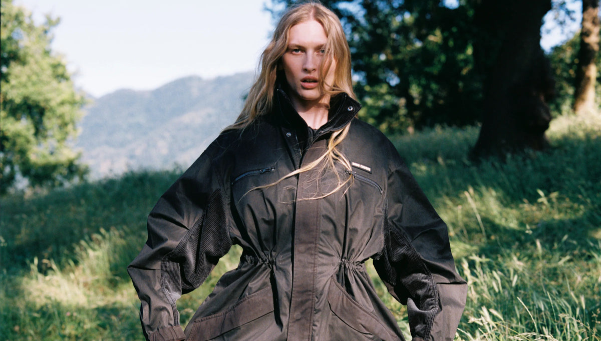 A woman stands outdoors in a grassy field with trees and hills in the background, wearing a dark oversized jacket with a drawstring waist and voluminous sleeves.