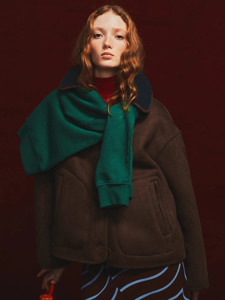 A model with red hair wearing a brown suede jacket, a dark green scarf, a red turtleneck, and patterned pants, set against a solid dark red background.