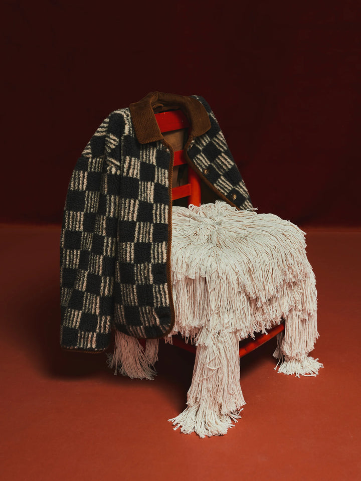 A styled product shot featuring a black and cream checkered knit jacket with a brown collar, draped over a red chair with a cream-colored shaggy fringe throw blanket.