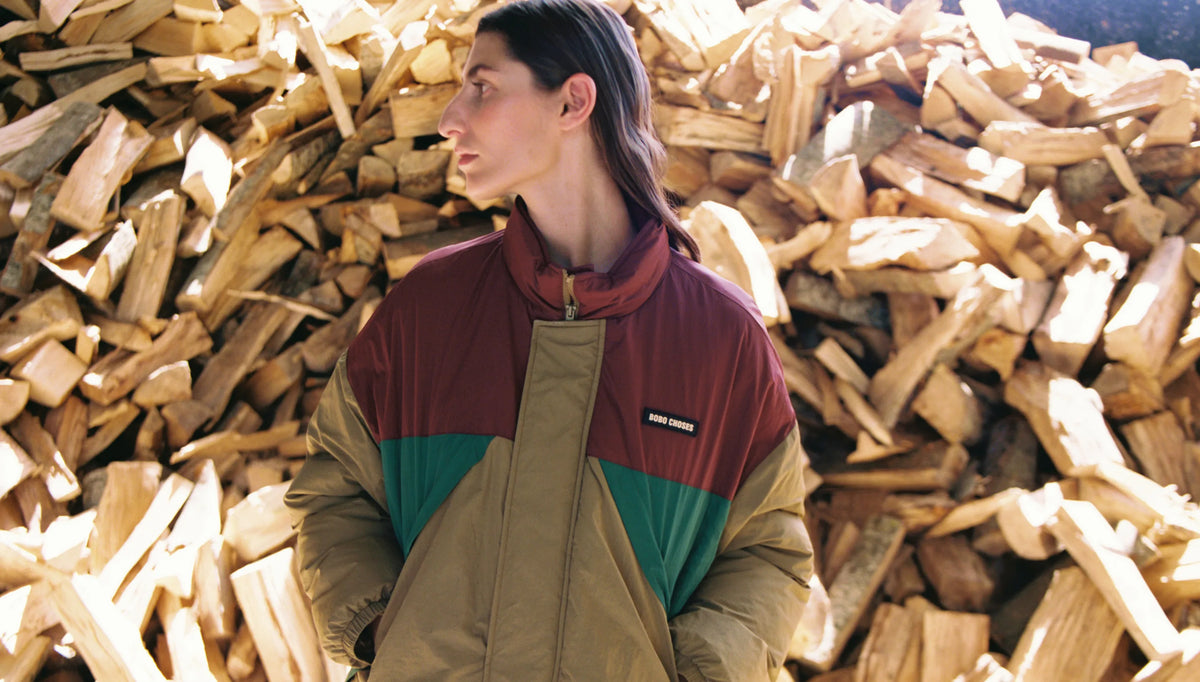 A woman wearing a Patagonia colorblock jacket stands in front of a large pile of cut firewood. The jacket features colorblocks of burgundy, green, and tan.