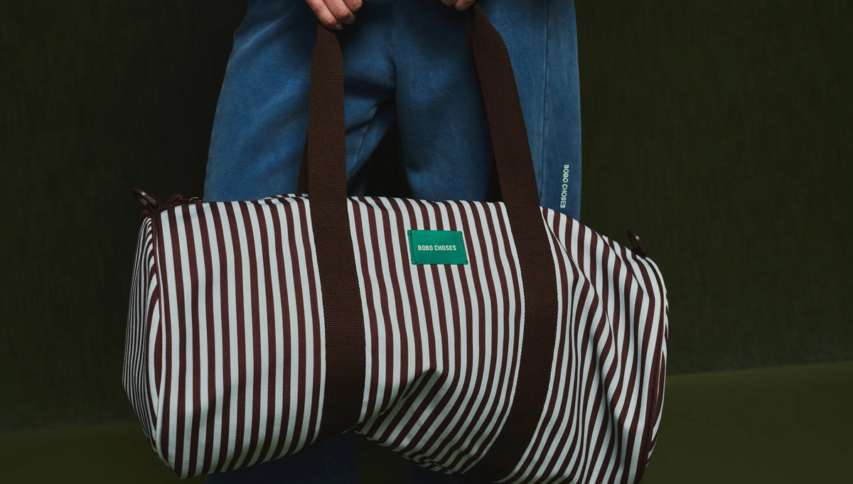 A striped duffel bag with brown handles and a green label, held by a person wearing blue jeans against a dark green background.