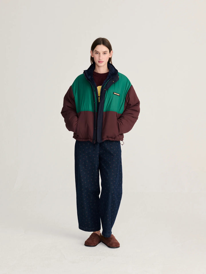 A model stands against a light gray studio background wearing a color-blocked puffer jacket with green and dark brown panels, dark patterned wide-leg pants, and brown shoes.