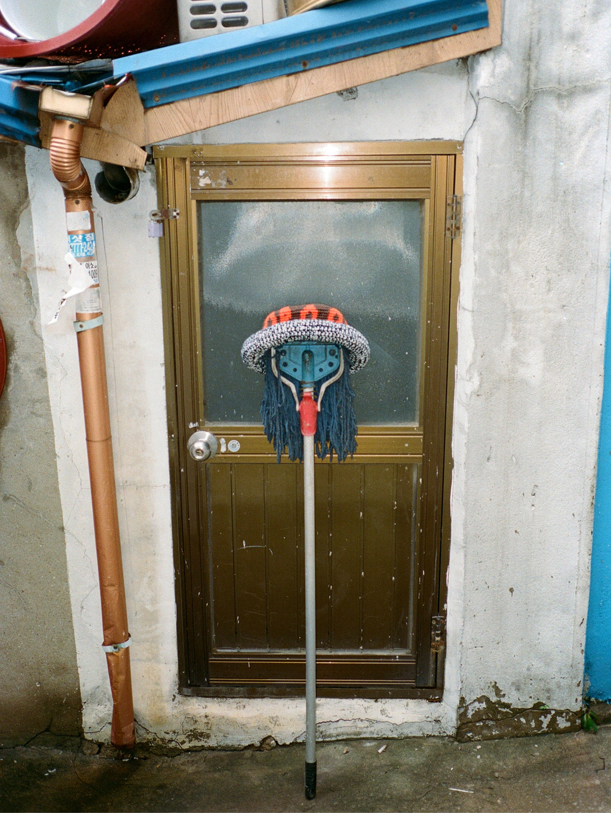 A long-handled floor mop with a blue and gray knit head and red handle, leaning in front of a brown door on a weathered concrete wall.