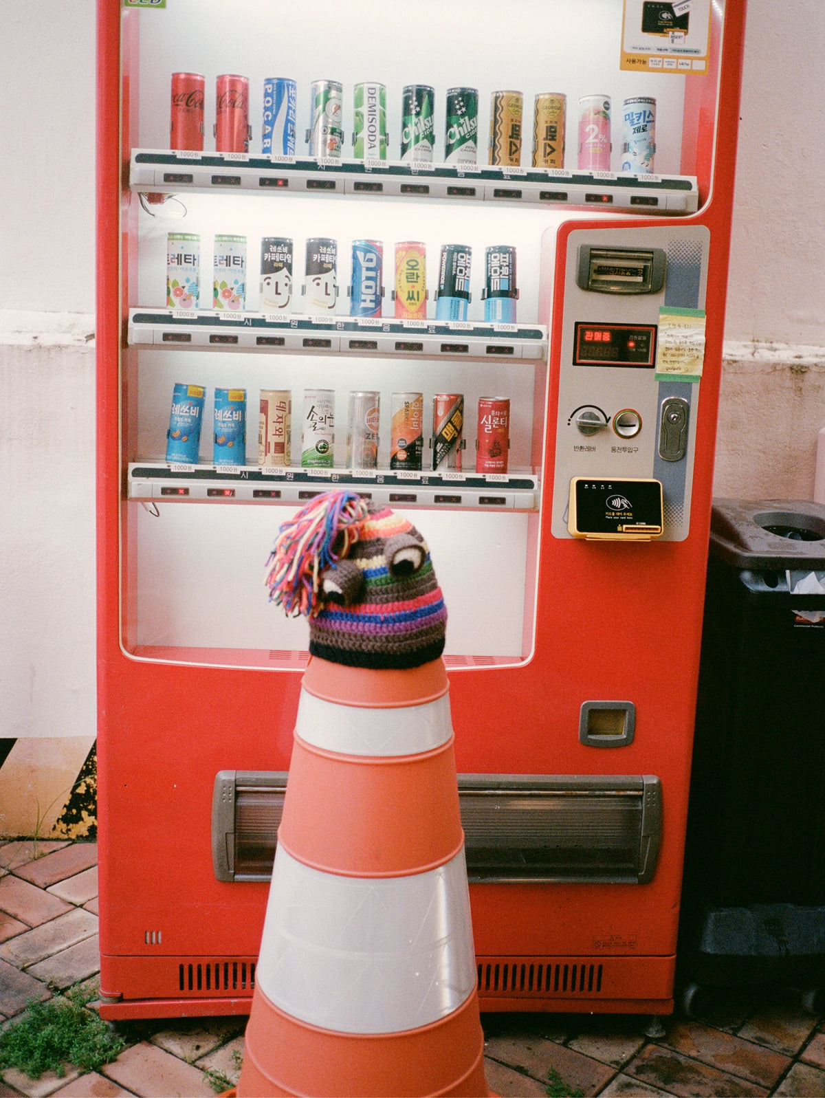 A red vending machine stands outdoors against a light wall, with a striped orange-and-white traffic cone in front holding a colorful knit hat with a small stuffed animal face.