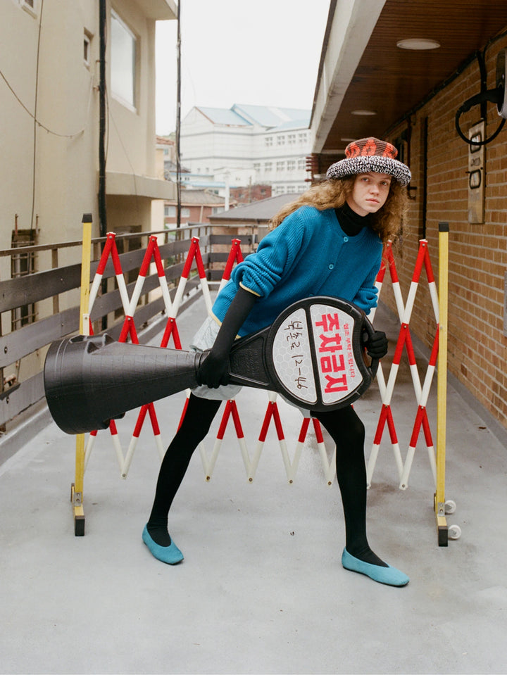 A model in a blue sweater, black tights, and a patterned hat holds a large black megaphone with a circular sign featuring Japanese text, set against a red and white collapsible barricade on an outdoor walkway.