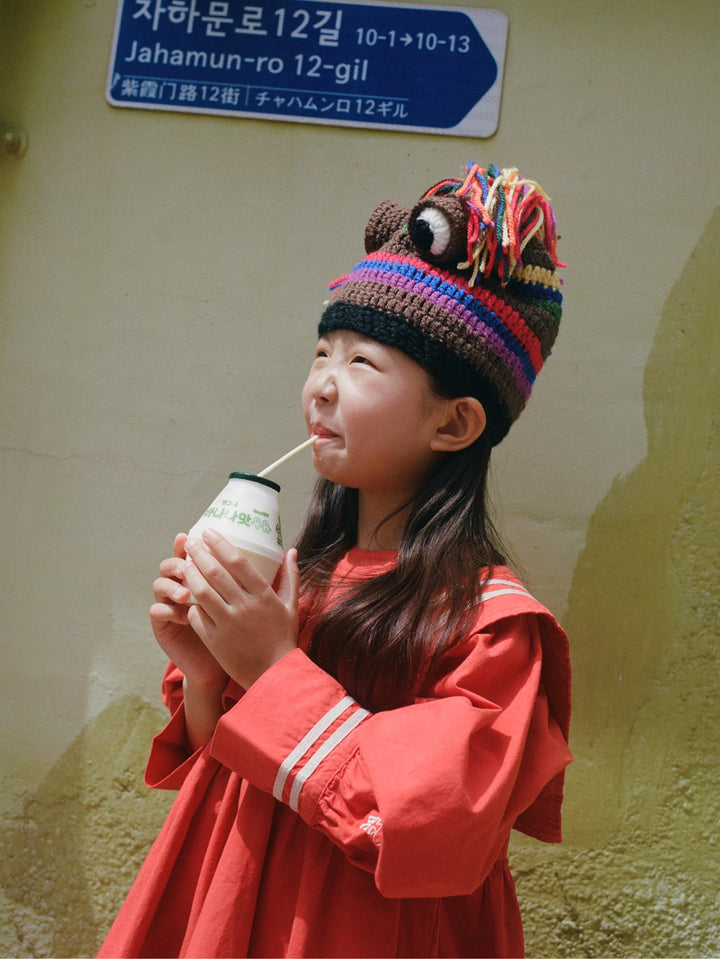 A young girl wearing a colorful striped knit beanie with a lion face and a red dress holds a milk bottle with a straw against a textured beige wall with a street sign in the background.