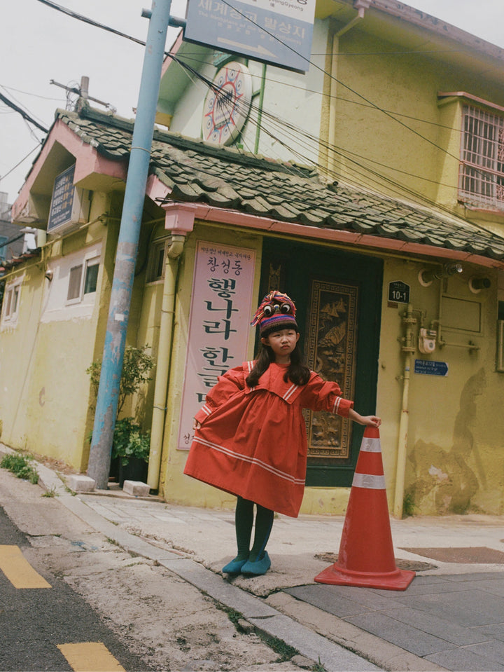 A young girl wearing a bright red traditional Korean dress and a colorful patterned hat stands on a street corner next to a yellow building, holding an orange traffic cone.