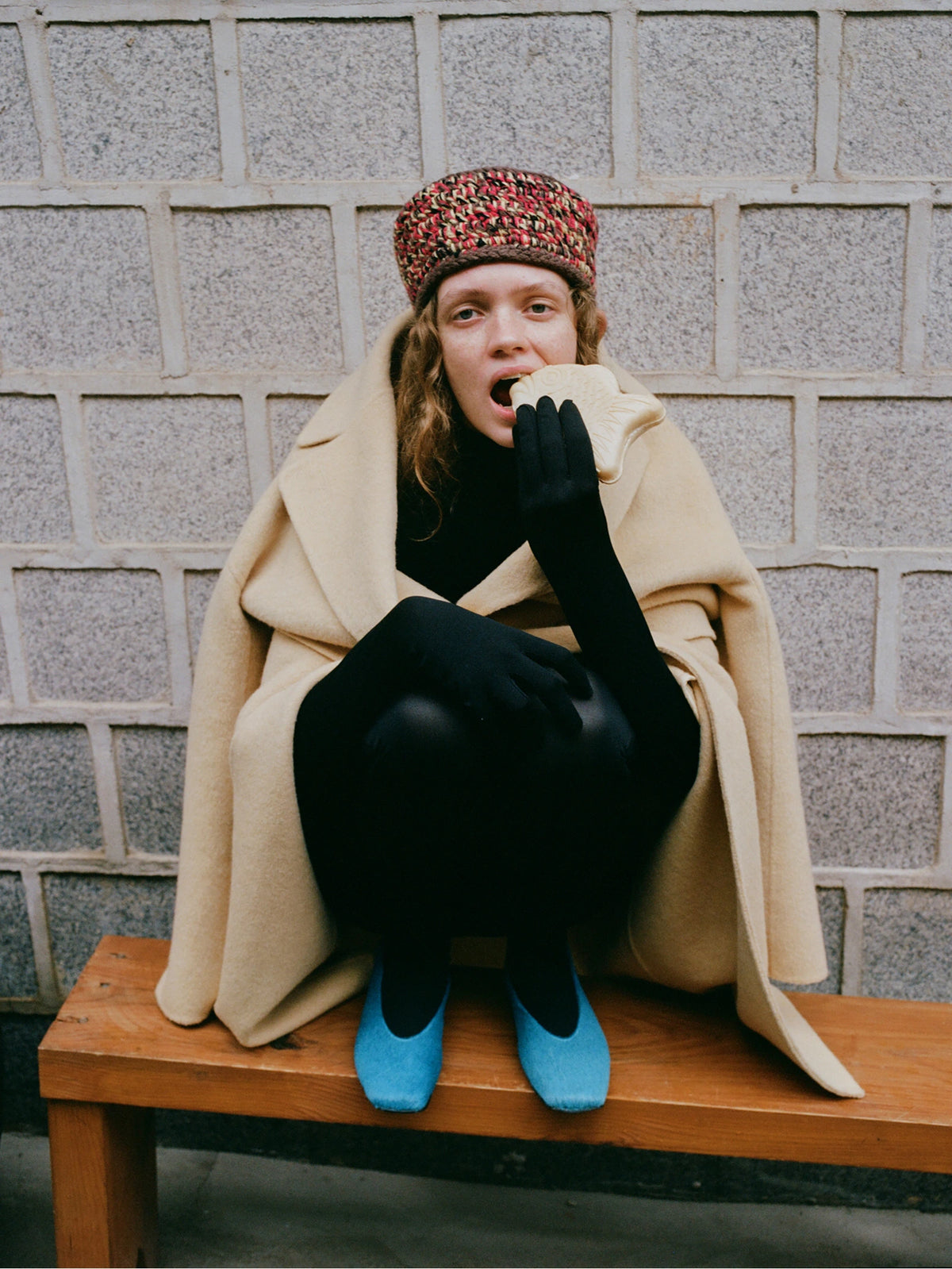 A model sits on a wooden bench against a gray stone wall, wrapped in a beige coat, wearing a patterned knit hat, black tights, and bright blue shoes.