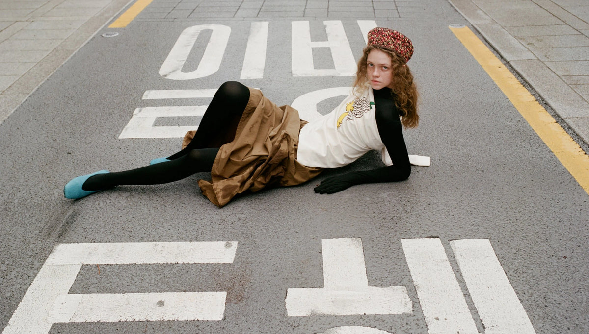 A model lies on an asphalt street crossing wearing a brown skirt, white graphic t-shirt, black long-sleeve top, black tights, and a red patterned beret.
