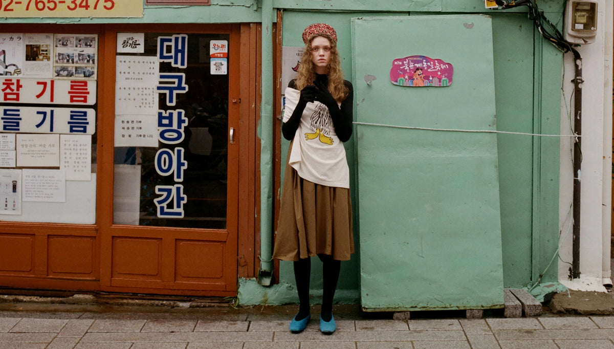 A person stands outdoors in front of a teal and orange storefront, wearing a patterned headband, black gloves, a cream graphic T-shirt, a brown skirt, black tights, and blue shoes.