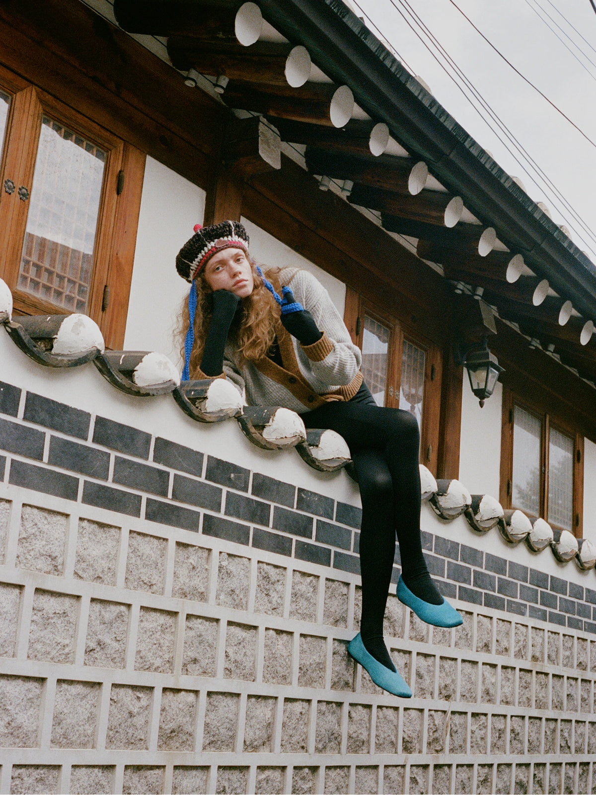 A woman wearing a gray knit sweater, black tights, a patterned knit hat, and bright turquoise slip-on shoes sits on the tiled edge of a traditional Korean house.