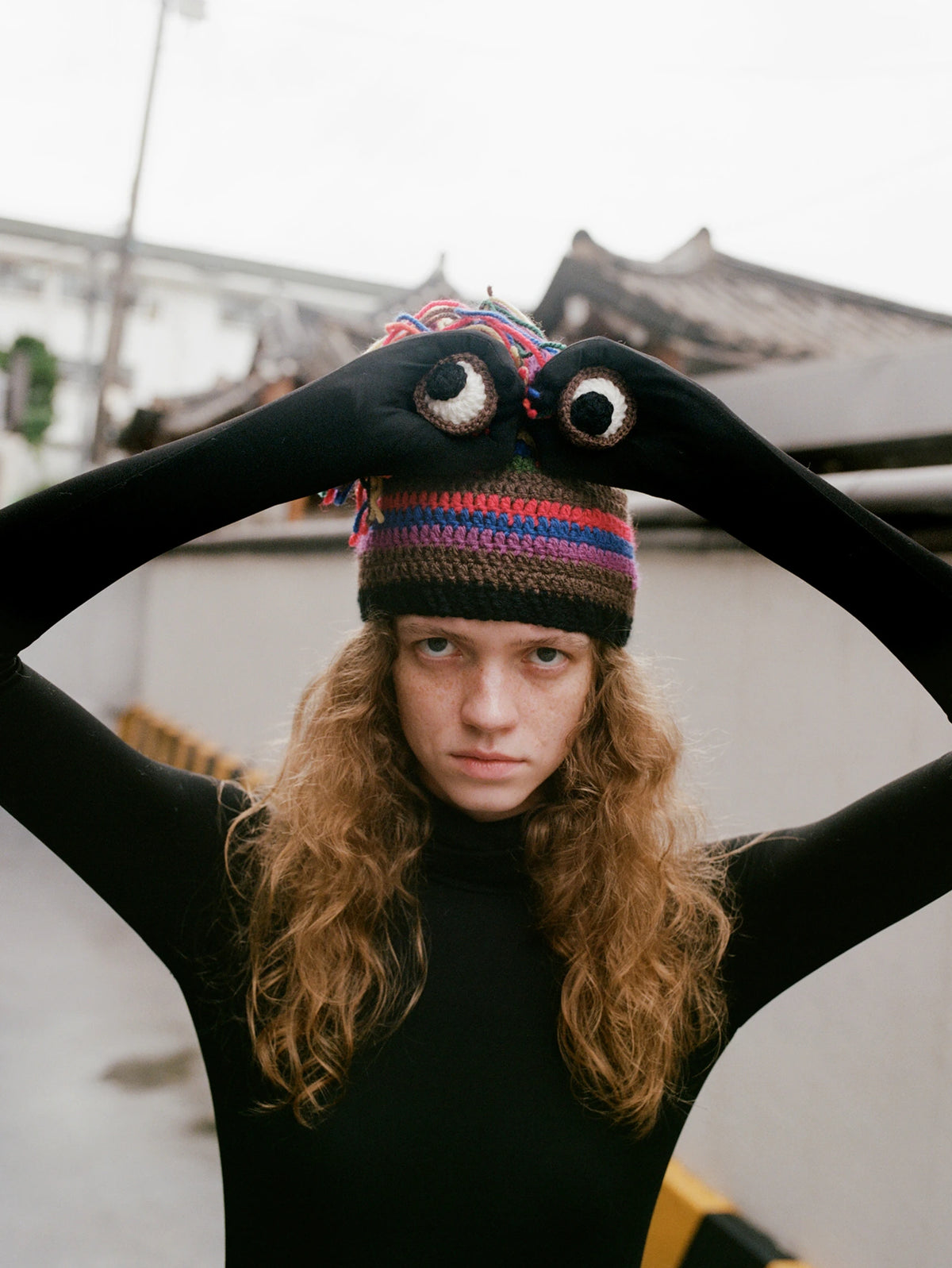 A person wearing a black long-sleeve top and a colorful striped knit beanie with large embroidered eyes, posing with arms raised against an outdoor background.
