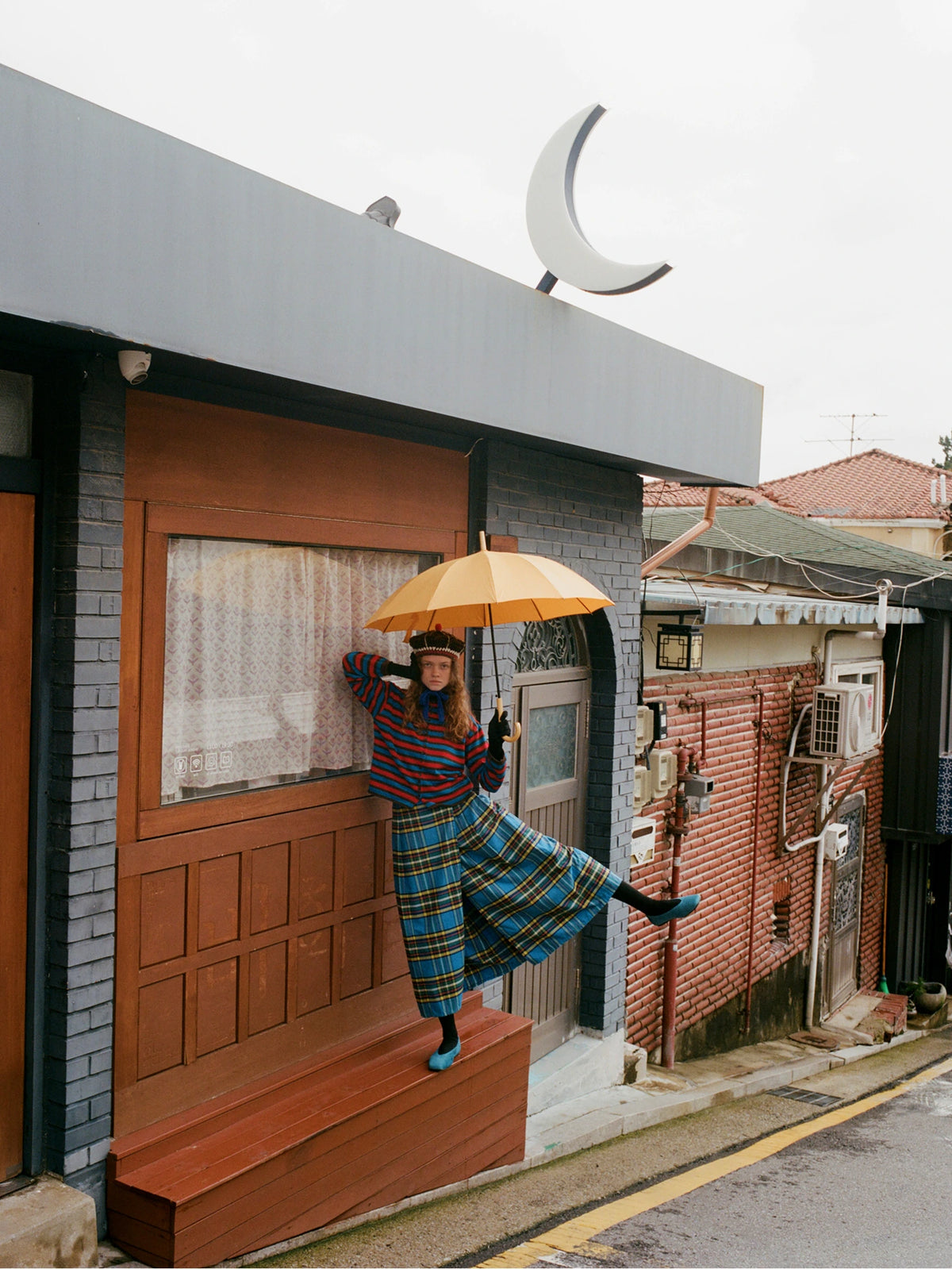 A person stands on a wooden step in front of a building, holding a yellow umbrella. They are wearing a colorful striped long-sleeve top and a blue, green, and yellow plaid skirt.