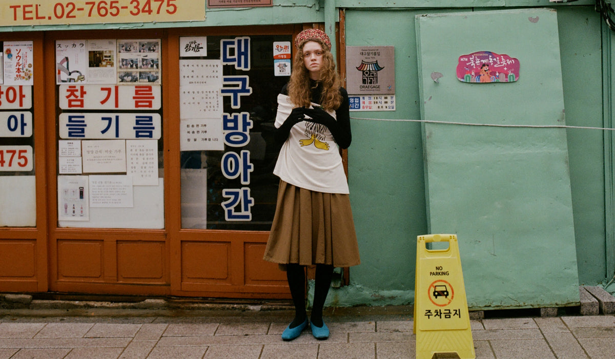 A woman stands in front of a storefront with Korean signage, wearing a brown hat, black top, white graphic T-shirt, brown pleated skirt, black tights, and blue shoes. A yellow caution sign is on the ground nearby.