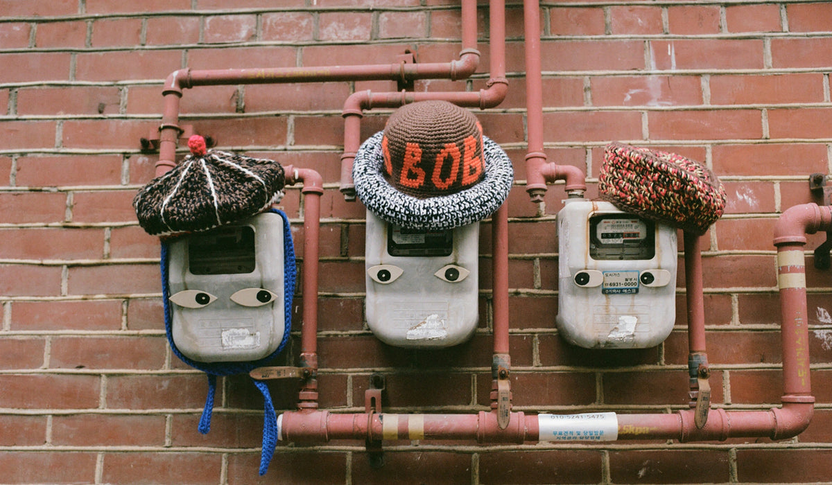 Three knitted winter hats are displayed on gray utility meters mounted on a red brick wall with exposed copper pipes. The center hat is brown and knitted with the word 'BOB' in orange, flanked by a dark patterned hat with a red pom-pom and a multicolor knit hat.