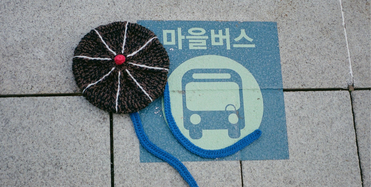 A handmade crocheted or knitted hat with a circular black crown, white stitched lines, and a red center, placed on a gray tiled surface next to a blue sign with Korean text and a bus icon.